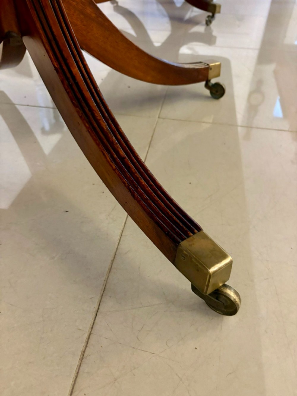 An extended mahogany dining table featuring a highly polished figured timber top supported by a three-pillar pedestal base with splayed legs and brass castors. View III