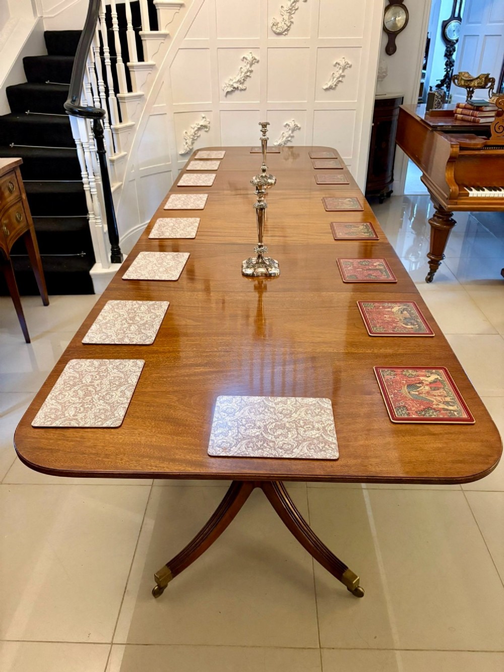 An extended mahogany dining table featuring a highly polished figured timber top supported by a three-pillar pedestal base with splayed legs and brass castors. View IV