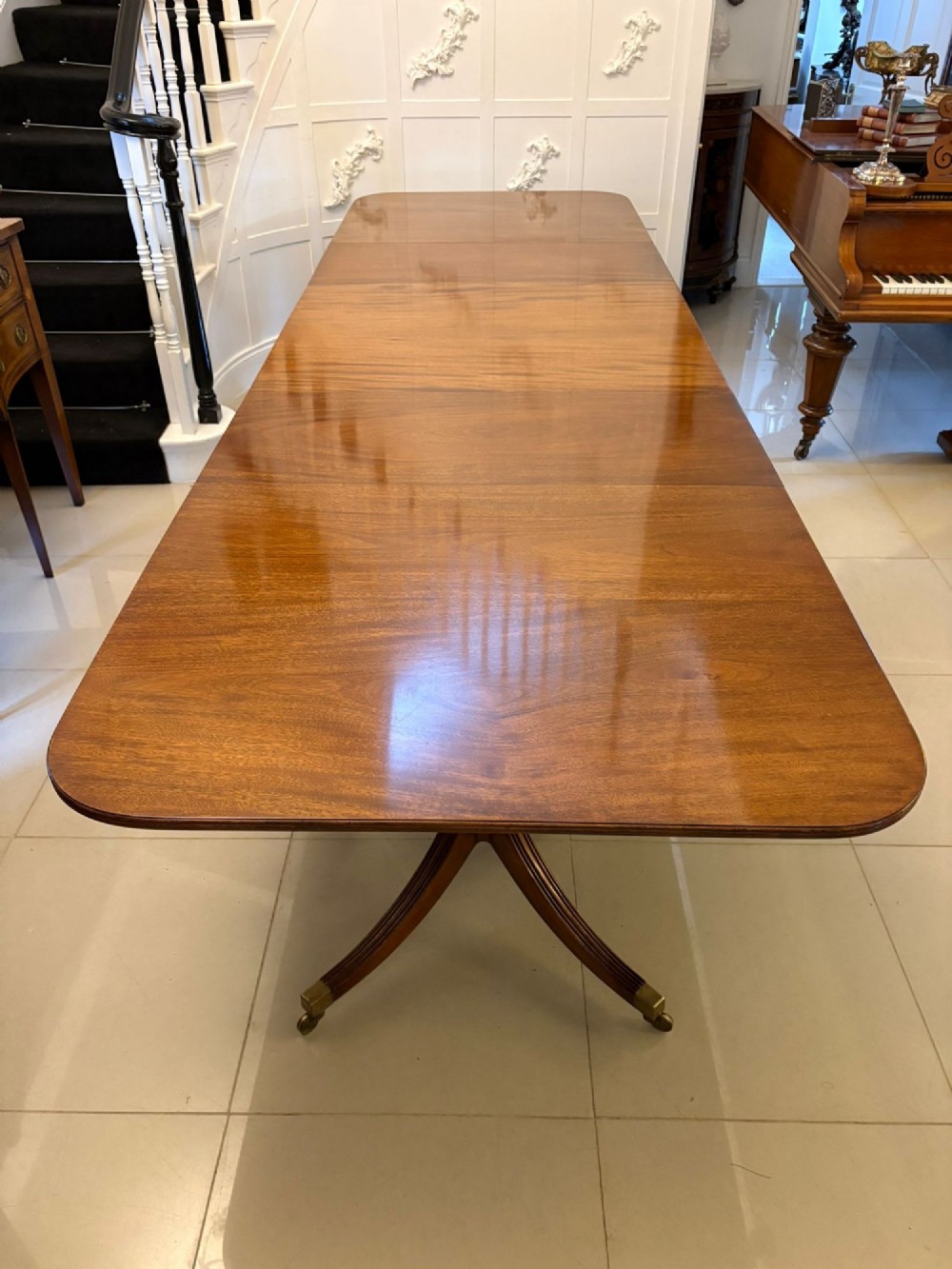 An extended mahogany dining table featuring a highly polished figured timber top supported by a three-pillar pedestal base with splayed legs and brass castors. View V