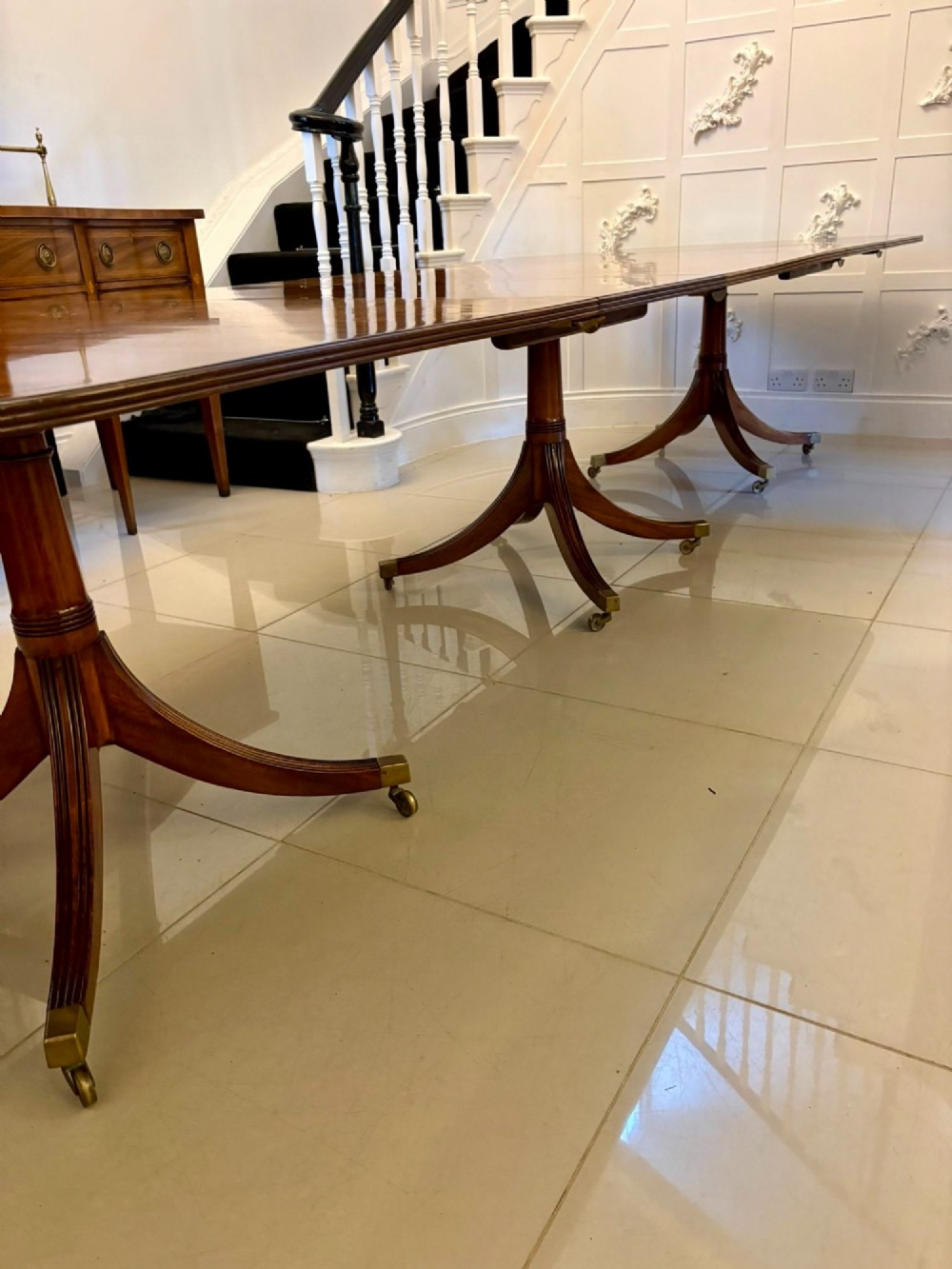 An extended mahogany dining table featuring a highly polished figured timber top supported by a three-pillar pedestal base with splayed legs and brass castors. View VI