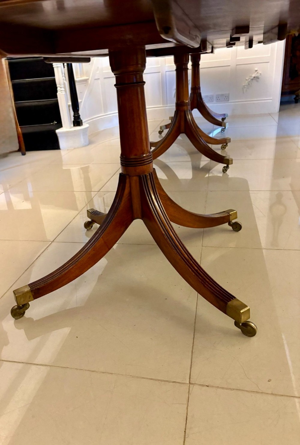 An extended mahogany dining table featuring a highly polished figured timber top supported by a three-pillar pedestal base with splayed legs and brass castors. View VII