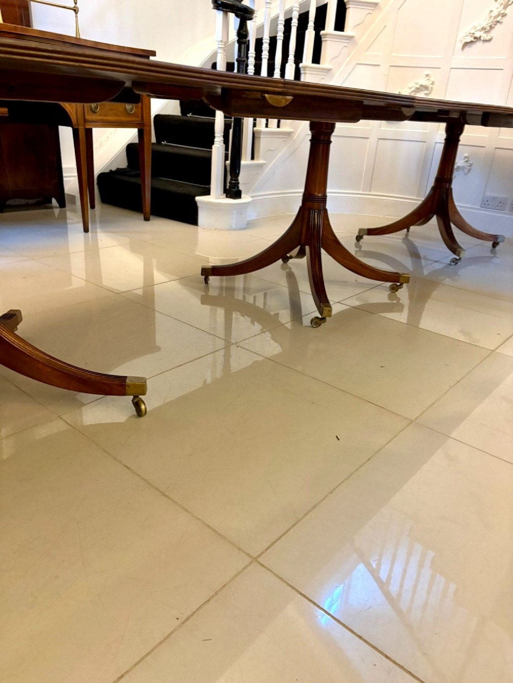 An extended mahogany dining table featuring a highly polished figured timber top supported by a three-pillar pedestal base with splayed legs and brass castors. View VIII