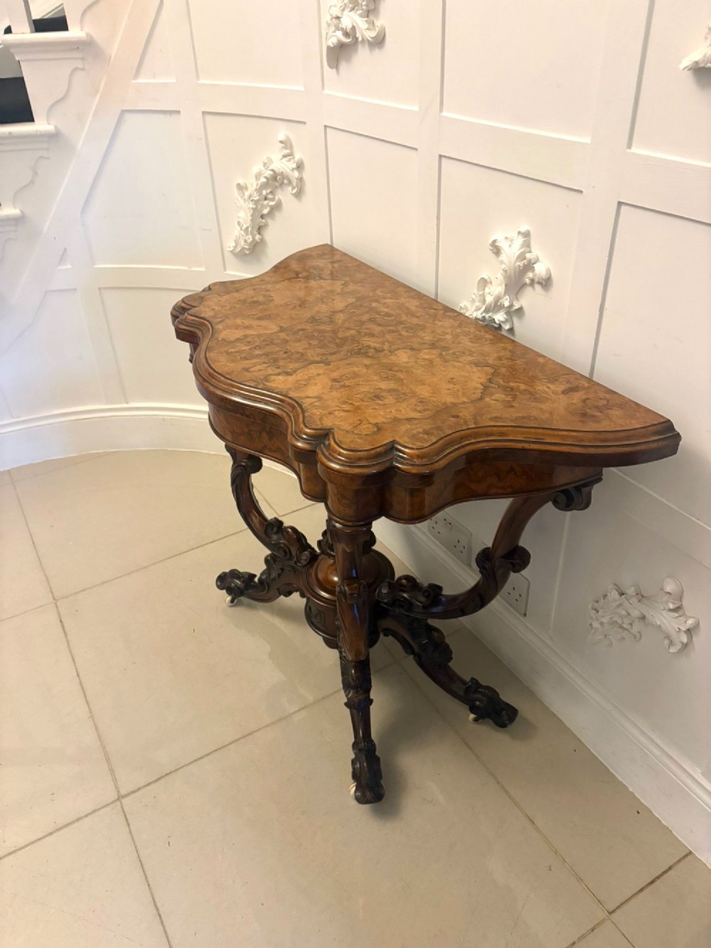 A Victorian burr walnut card table featuring a serpentine-shaped fold-over top set upon a carved pedestal base with four scroll feet on castors. View X