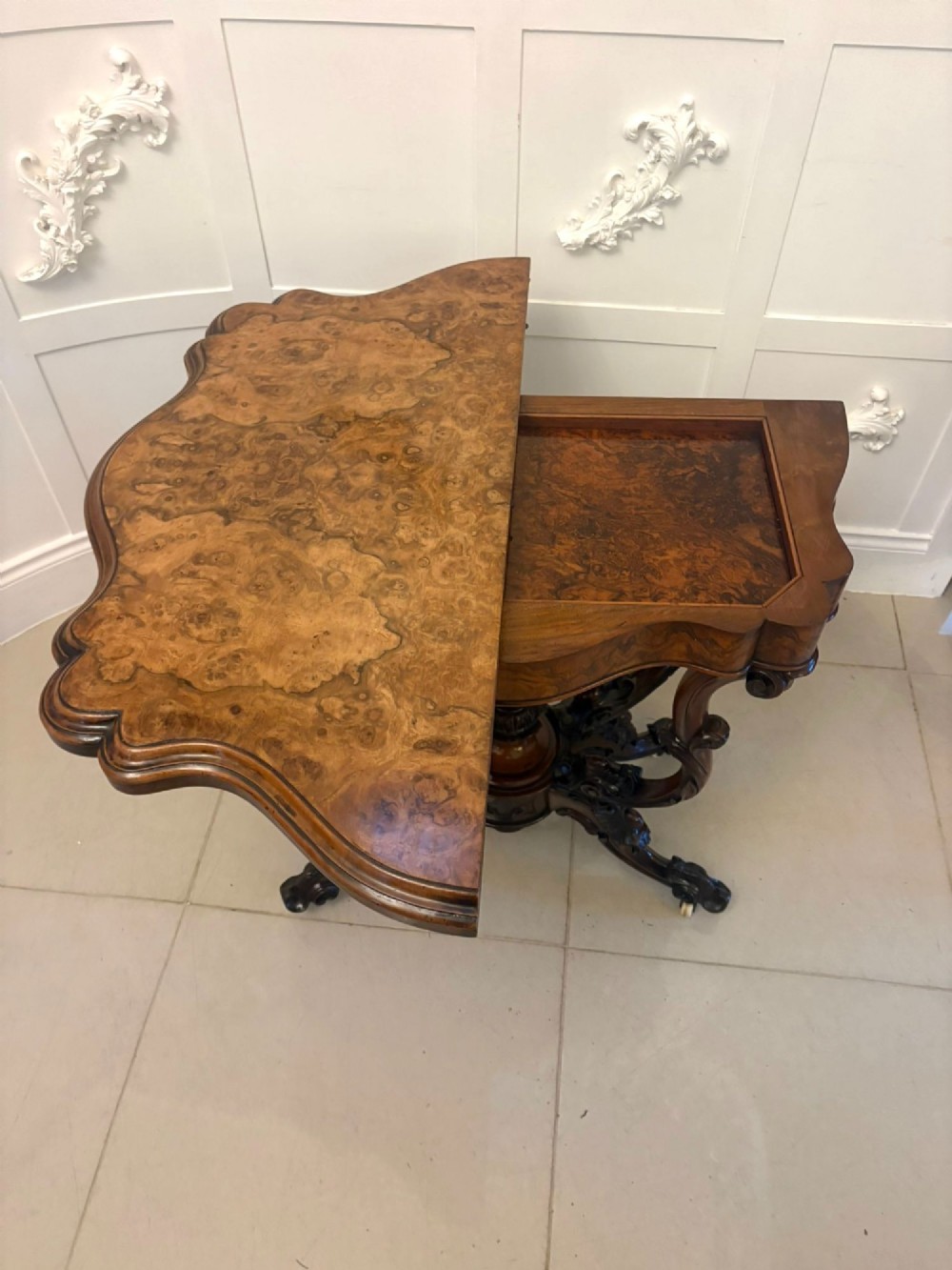 A Victorian burr walnut card table featuring a serpentine-shaped fold-over top set upon a carved pedestal base with four scroll feet on castors. View XII