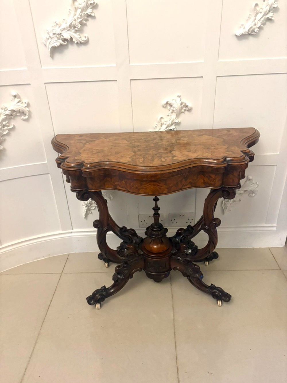 A Victorian burr walnut card table featuring a serpentine-shaped fold-over top set upon a carved pedestal base with four scroll feet on castors.