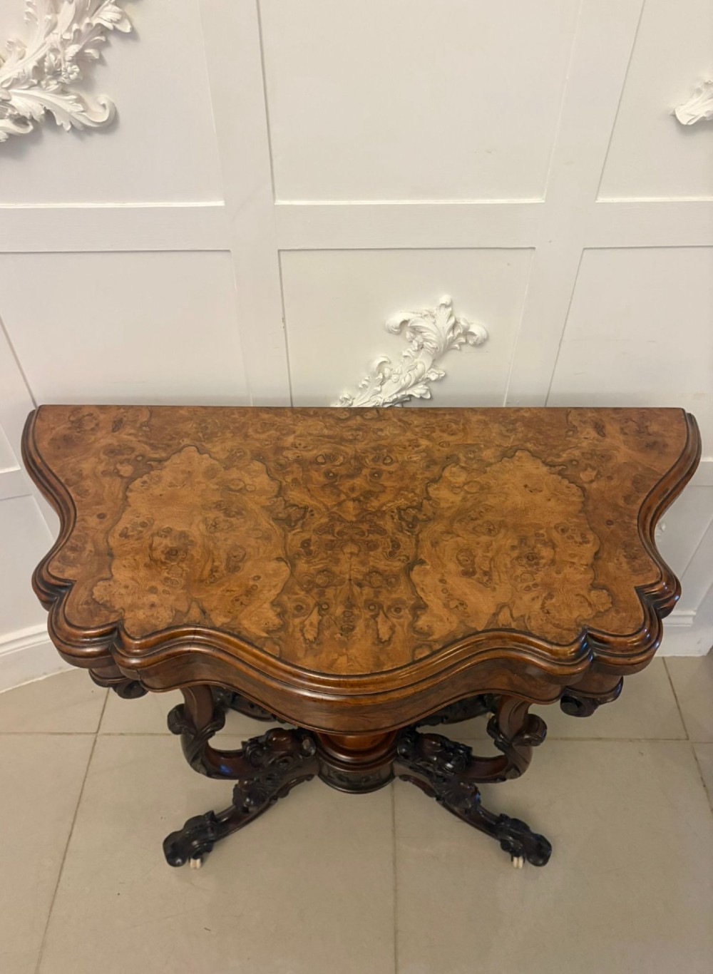 A Victorian burr walnut card table featuring a serpentine-shaped fold-over top set upon a carved pedestal base with four scroll feet on castors. View XV