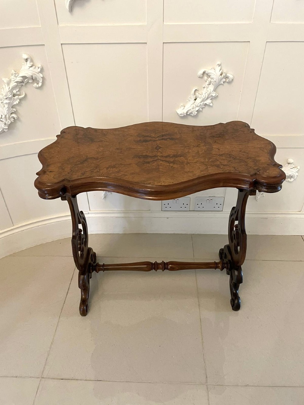 A Victorian burr walnut centre table with a serpentine shaped top, resting on carved pierced end supports and connected by a turned stretcher. View II