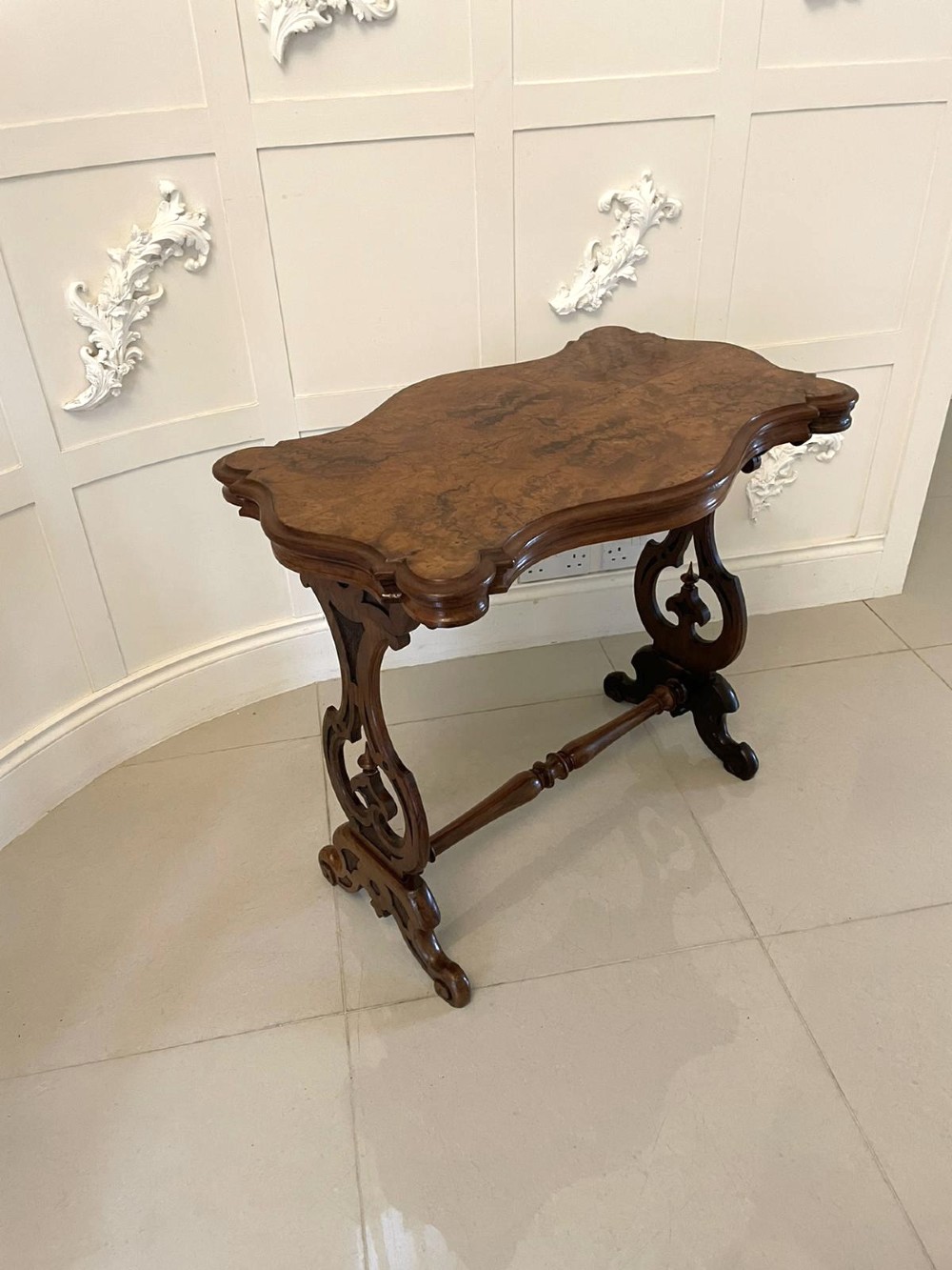 A Victorian burr walnut centre table with a serpentine shaped top, resting on carved pierced end supports and connected by a turned stretcher. View III
