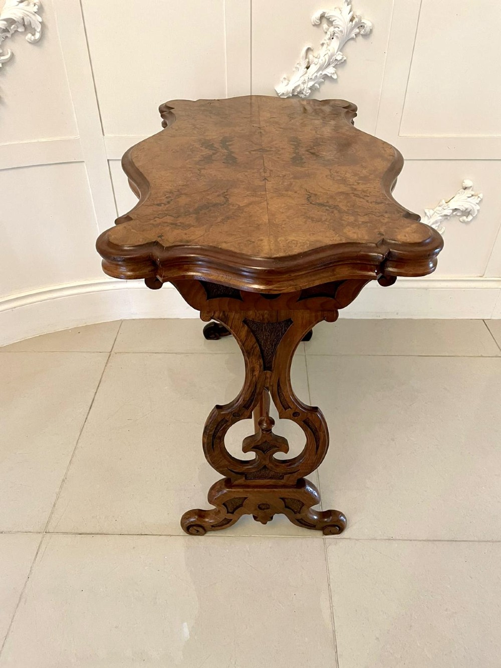 A Victorian burr walnut centre table with a serpentine shaped top, resting on carved pierced end supports and connected by a turned stretcher. View IV