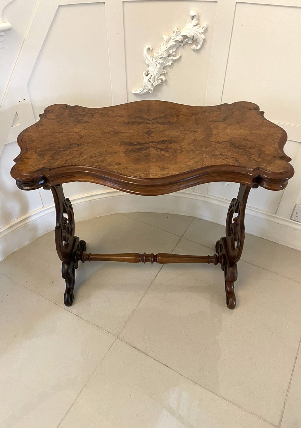 A Victorian burr walnut centre table with a serpentine shaped top, resting on carved pierced end supports and connected by a turned stretcher. View IX