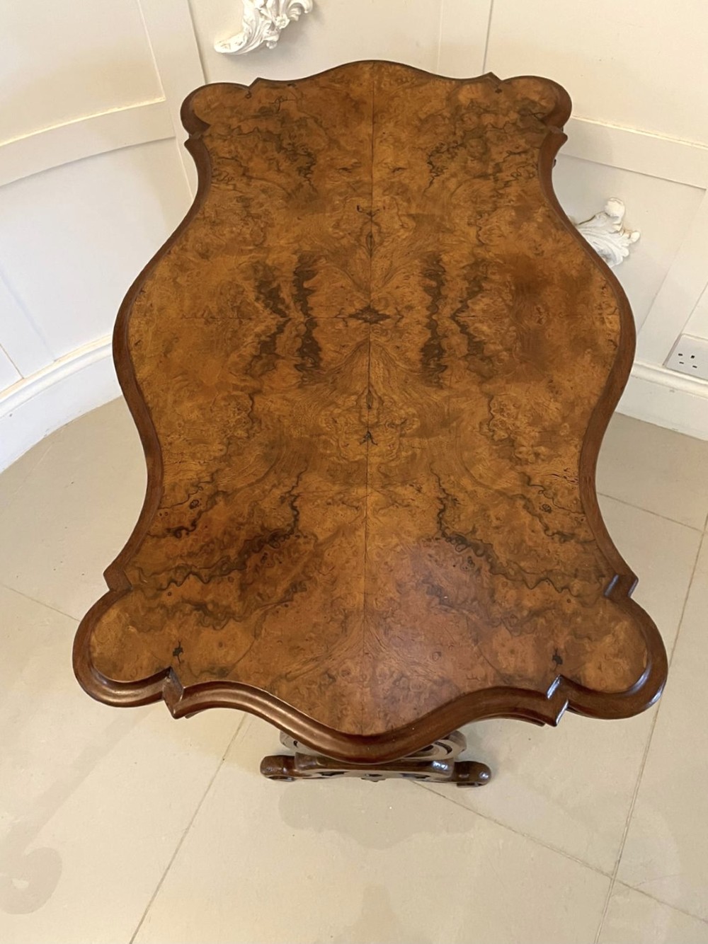 A Victorian burr walnut centre table with a serpentine shaped top, resting on carved pierced end supports and connected by a turned stretcher. View XI