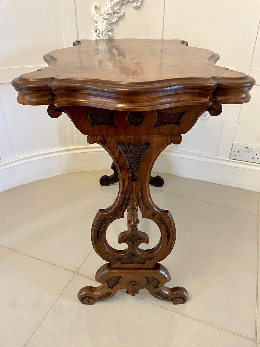 A Victorian burr walnut centre table with a serpentine shaped top, resting on carved pierced end supports and connected by a turned stretcher. View XII