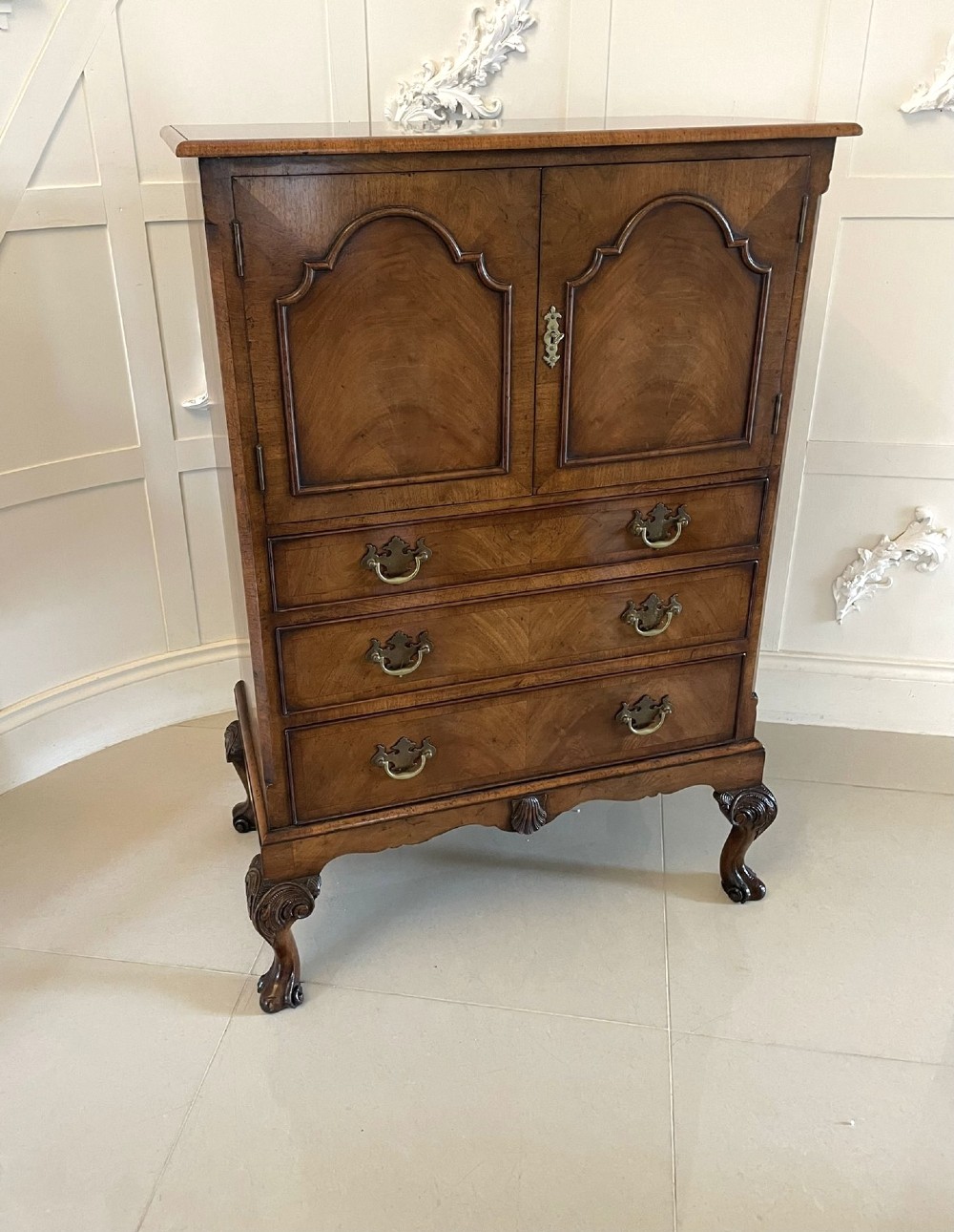 An antique burr walnut chest on stand featuring a two-door cupboard with arched panels above three long drawers, resting on carved cabriole legs.