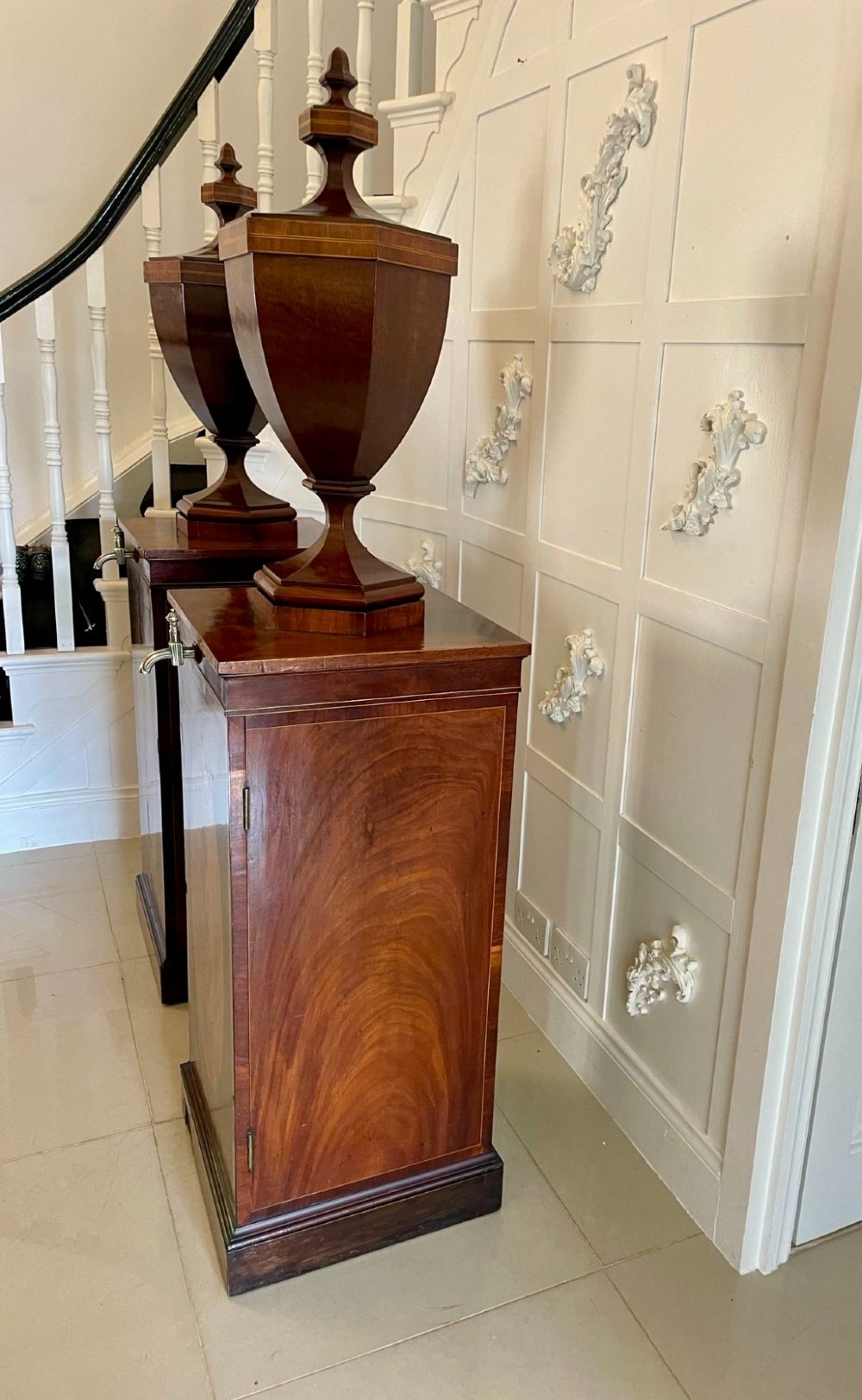 A pair of George III mahogany wine urns featuring flame mahogany oval inlay panels resting atop matching square pedestal cupboards with brass spigots. View IX
