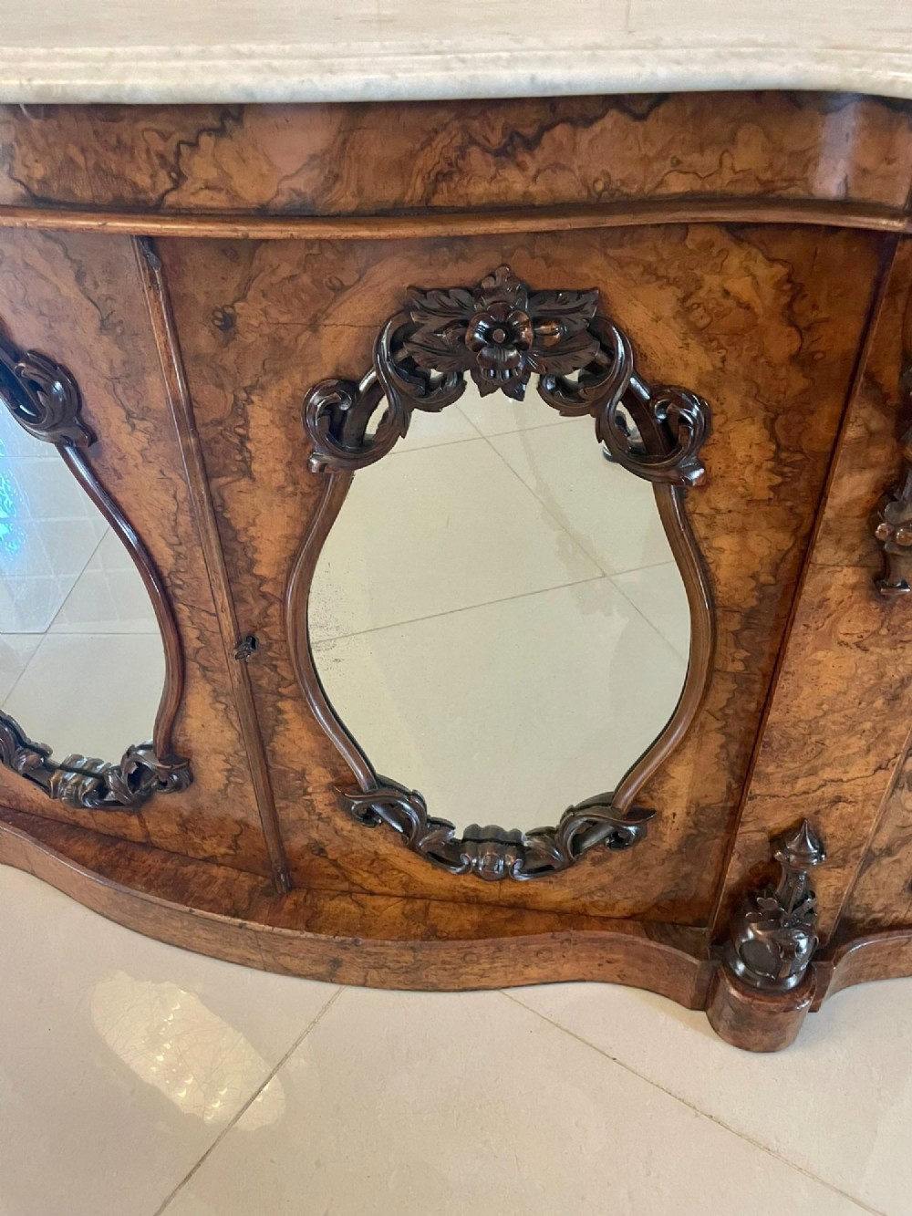 A Victorian burr walnut credenza featuring a serpentine white marble top, mirrored cupboard doors, and a large oval mirror back with carved scrolling detail. View XIV