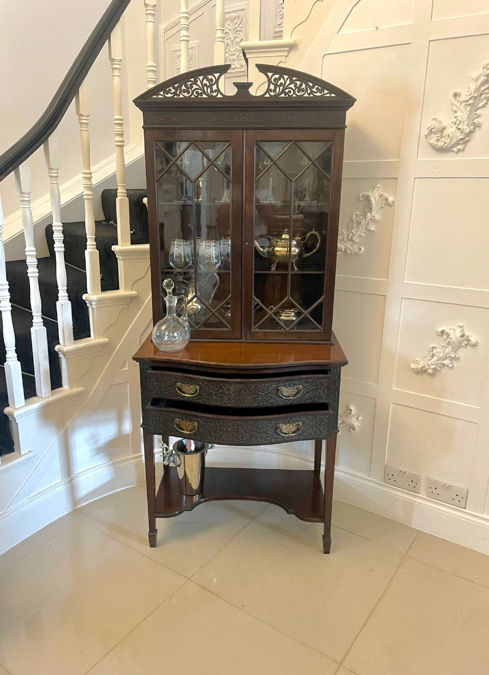 A dark carved mahogany display cabinet featuring astragal glazed doors, a pierced scrolled pediment, serpentine drawer fronts with brass handles, and a lower shelf. View III
