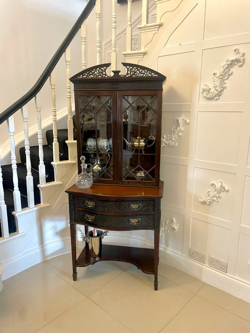 A dark carved mahogany display cabinet featuring astragal glazed doors, a pierced scrolled pediment, serpentine drawer fronts with brass handles, and a lower shelf. View IV