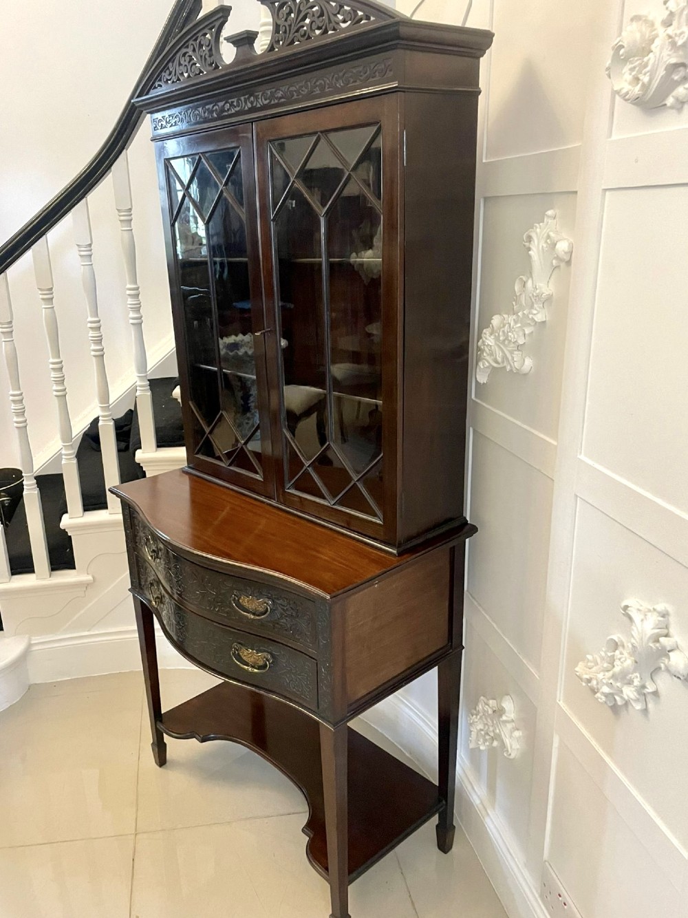 A dark carved mahogany display cabinet featuring astragal glazed doors, a pierced scrolled pediment, serpentine drawer fronts with brass handles, and a lower shelf. View V