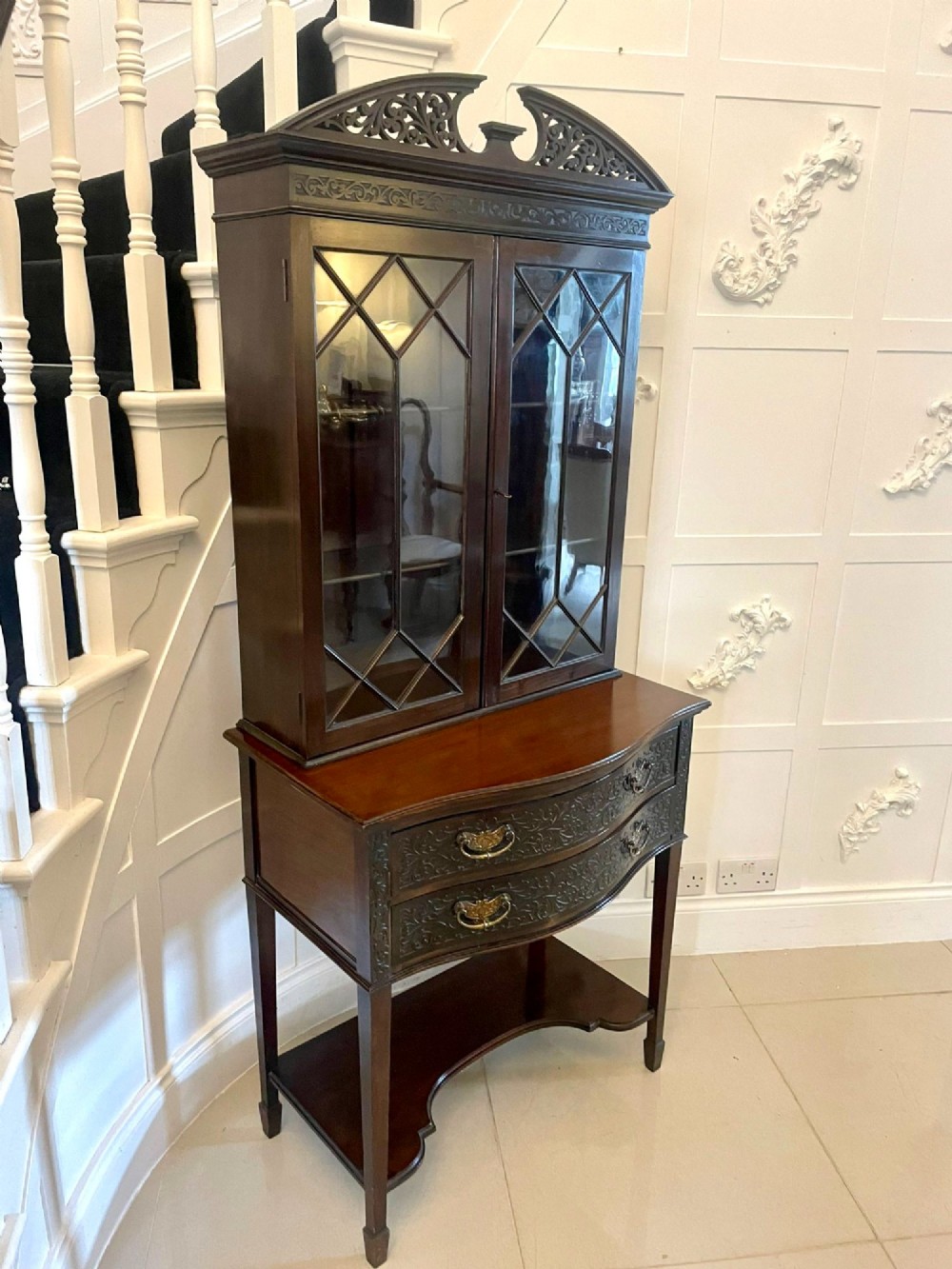 A dark carved mahogany display cabinet featuring astragal glazed doors, a pierced scrolled pediment, serpentine drawer fronts with brass handles, and a lower shelf. View VI
