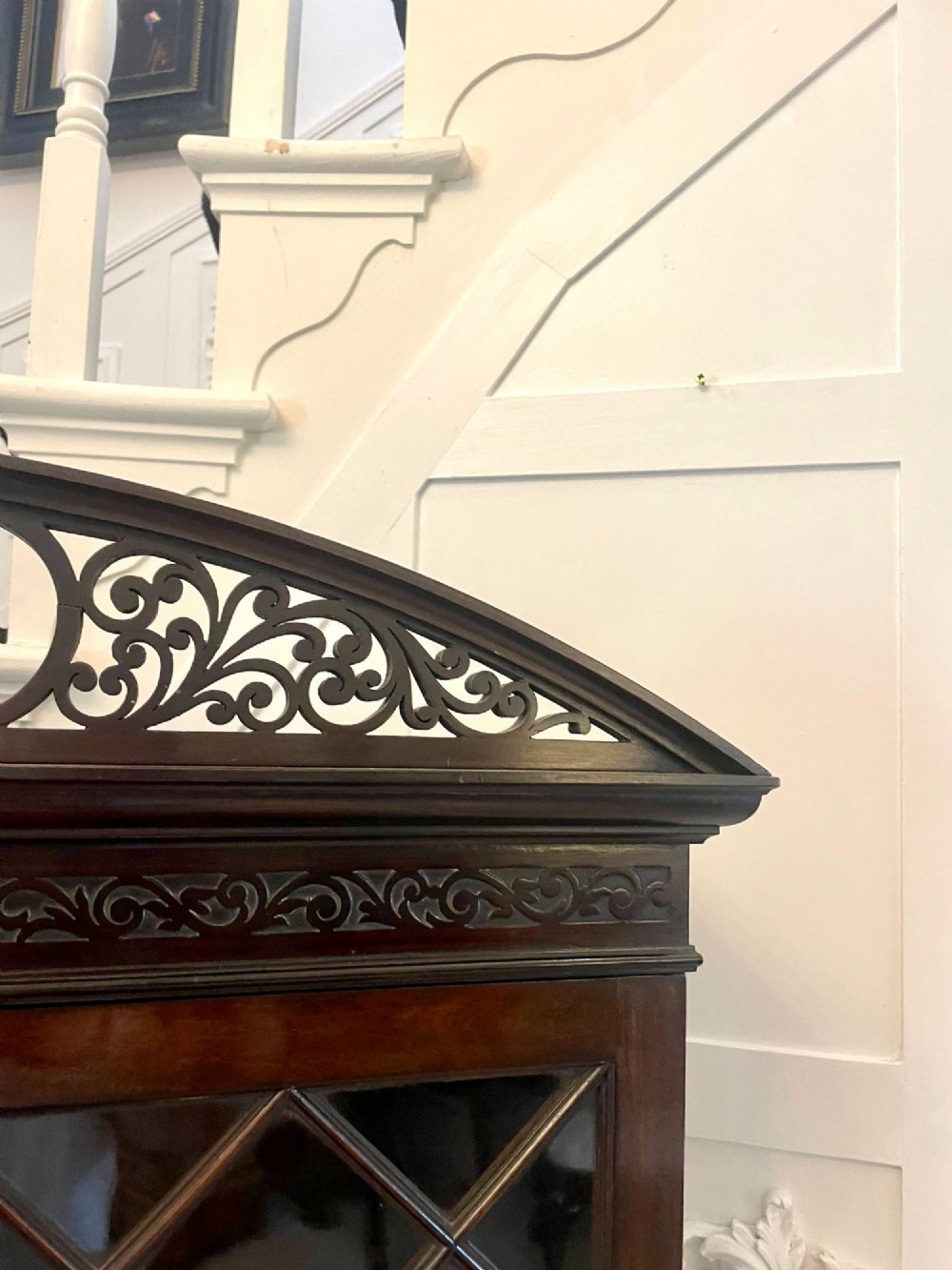 A dark carved mahogany display cabinet featuring astragal glazed doors, a pierced scrolled pediment, serpentine drawer fronts with brass handles, and a lower shelf. View VII