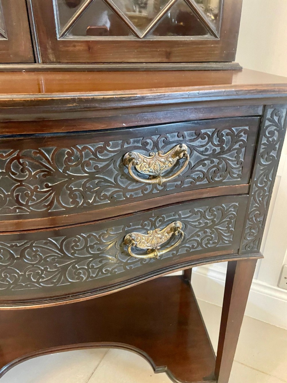 A dark carved mahogany display cabinet featuring astragal glazed doors, a pierced scrolled pediment, serpentine drawer fronts with brass handles, and a lower shelf. View X