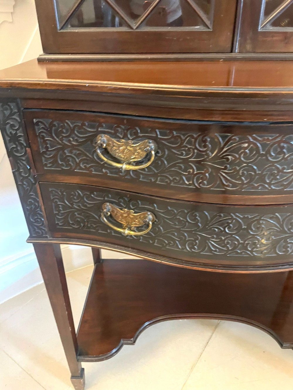 A dark carved mahogany display cabinet featuring astragal glazed doors, a pierced scrolled pediment, serpentine drawer fronts with brass handles, and a lower shelf. View XI