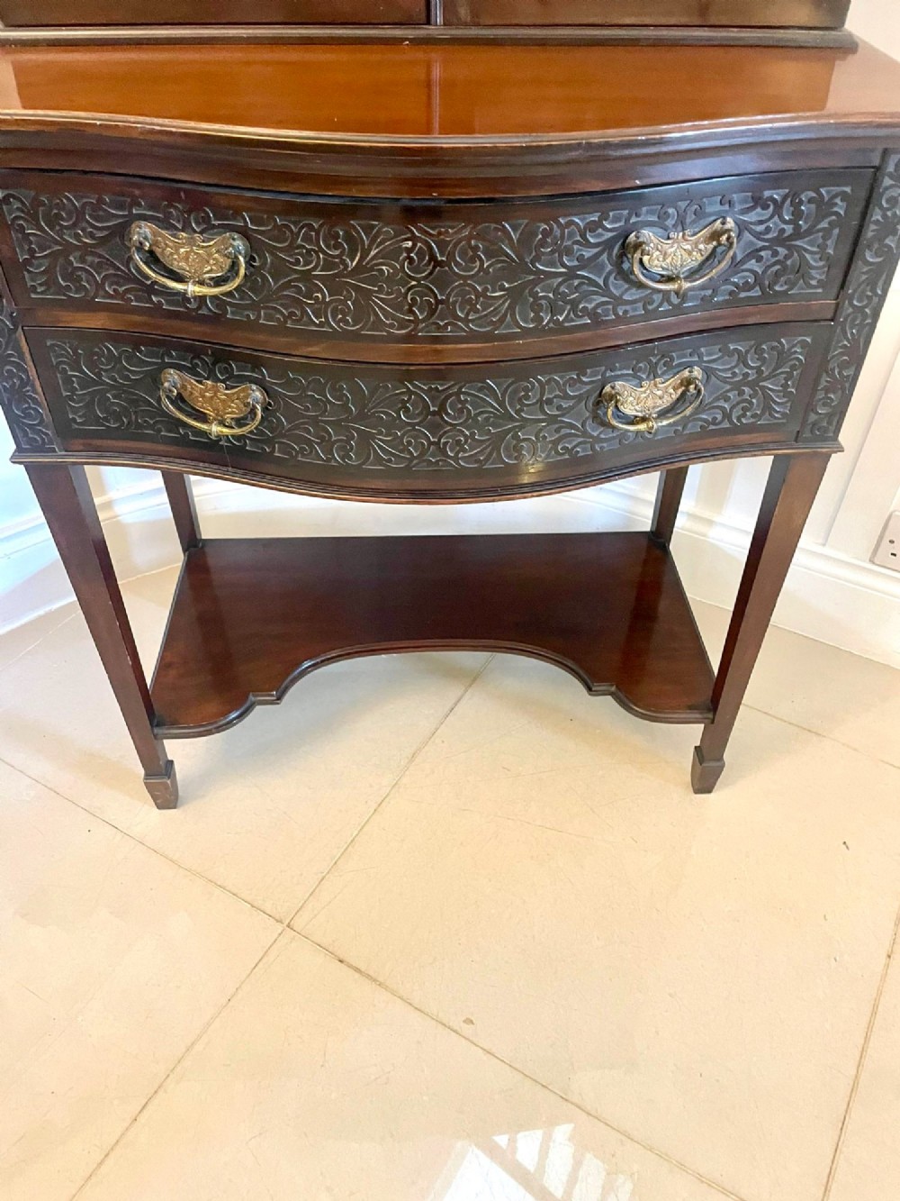 A dark carved mahogany display cabinet featuring astragal glazed doors, a pierced scrolled pediment, serpentine drawer fronts with brass handles, and a lower shelf. View XII