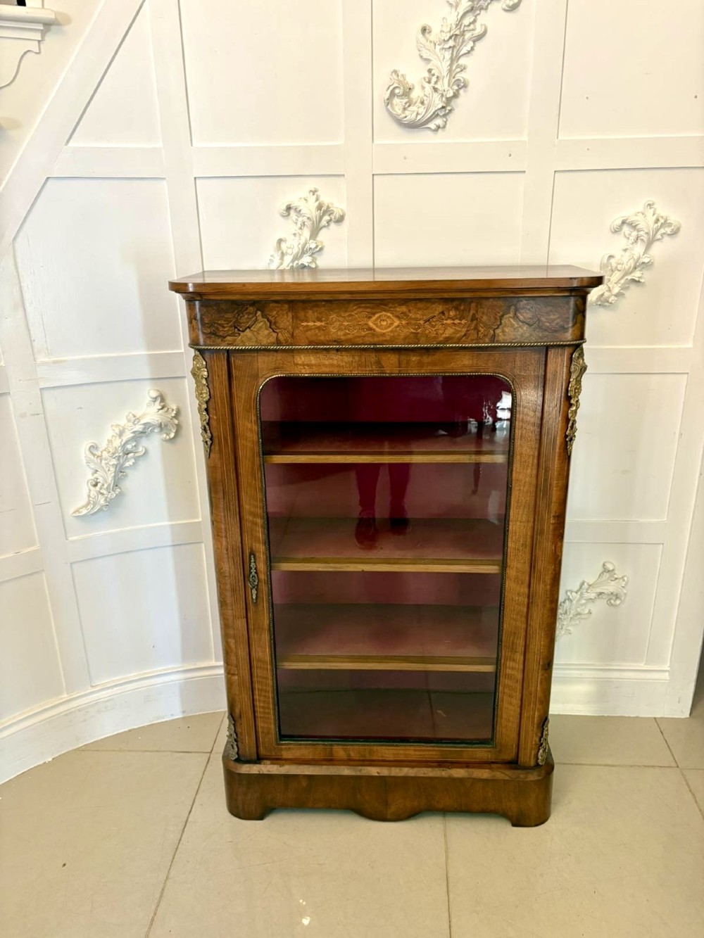 A Victorian burr walnut pier cabinet featuring a single glazed door, floral marquetry along the frieze, and gilt ormolu corner mounts.