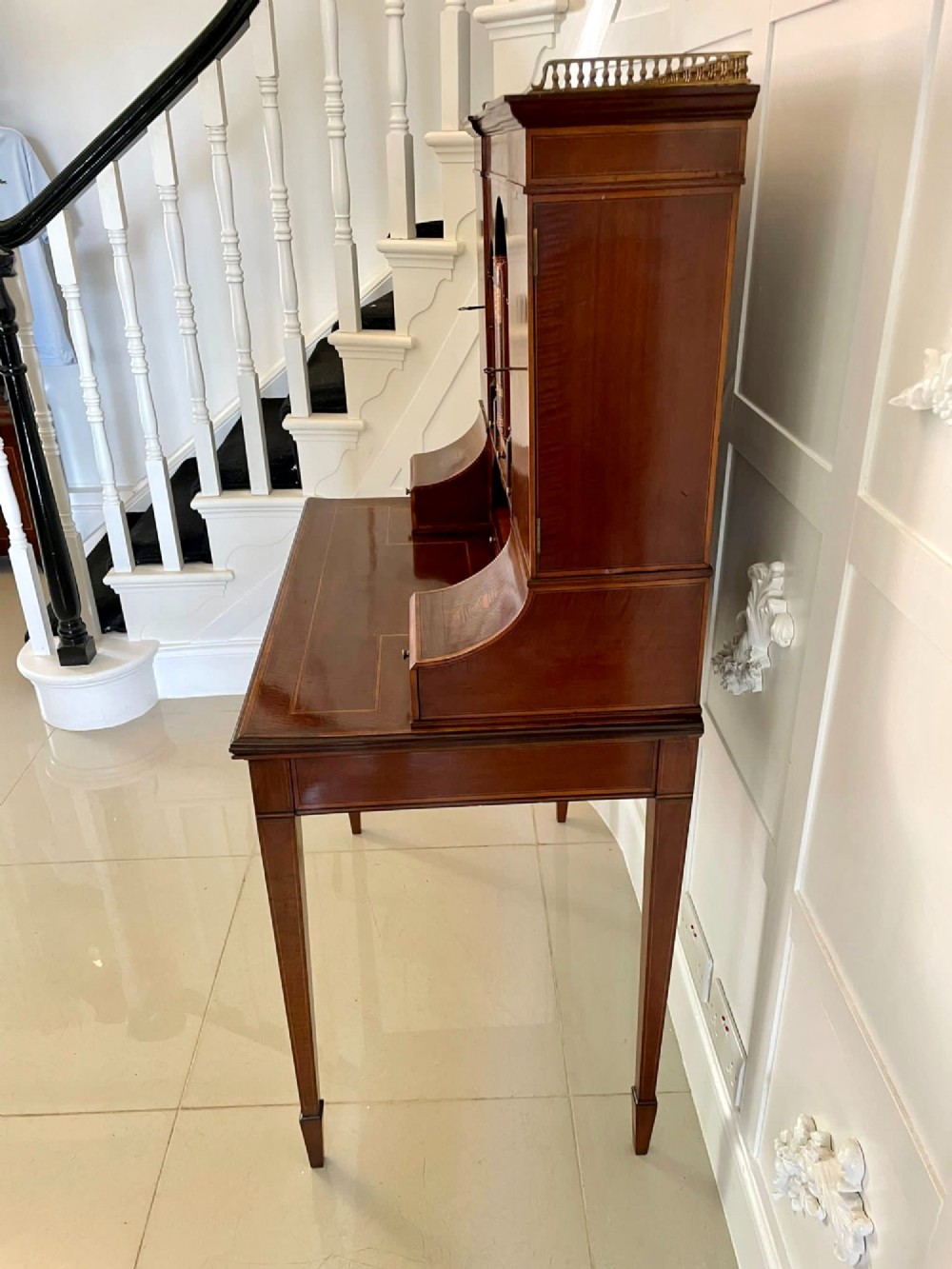 A Victorian inlaid mahogany writing desk featuring a brass gallery top, mirrored arched niche, floral marquetry cabinets, small drawers, and tapered square legs. View VI