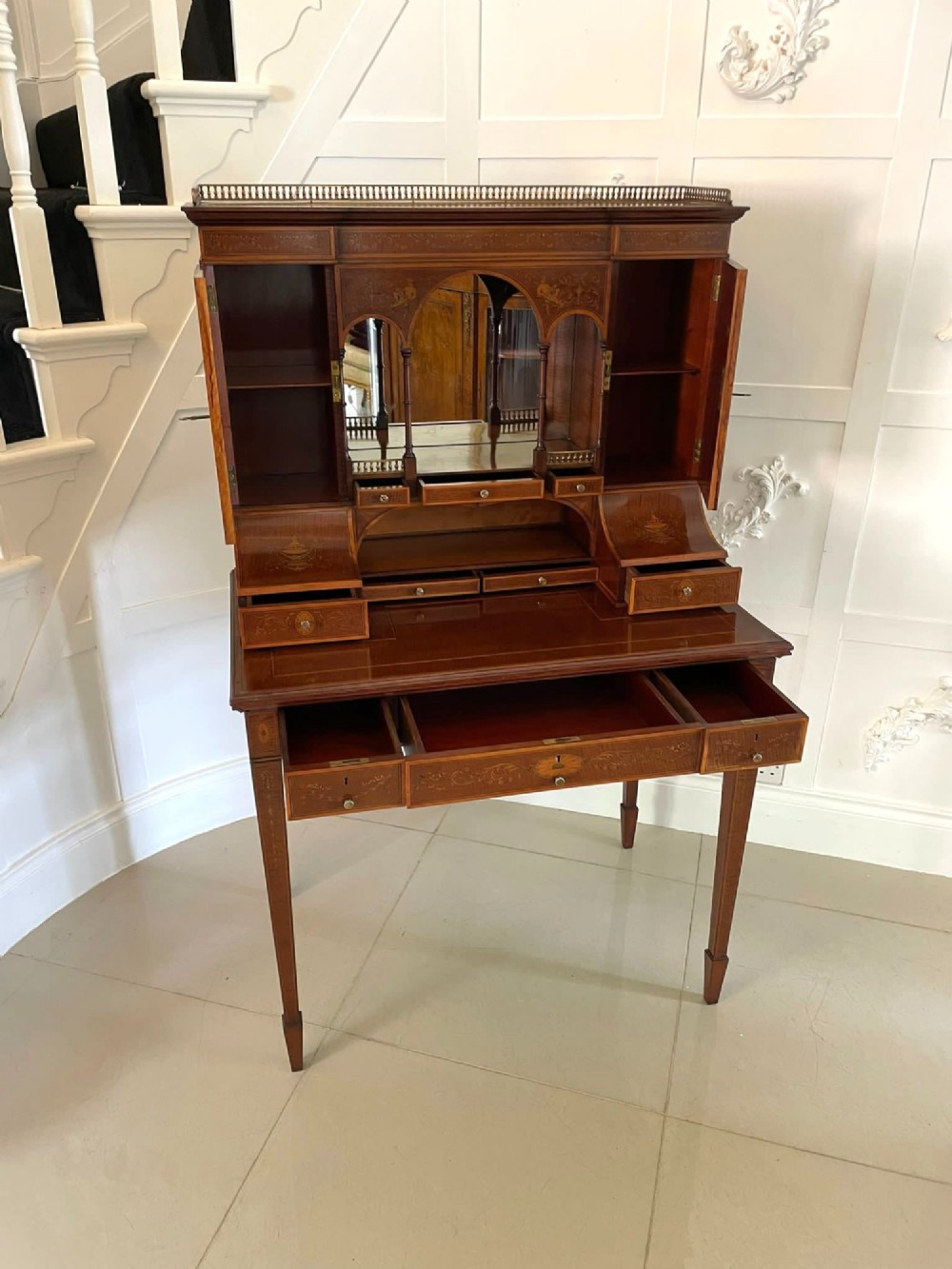 A Victorian inlaid mahogany writing desk featuring a brass gallery top, mirrored arched niche, floral marquetry cabinets, small drawers, and tapered square legs. View XI