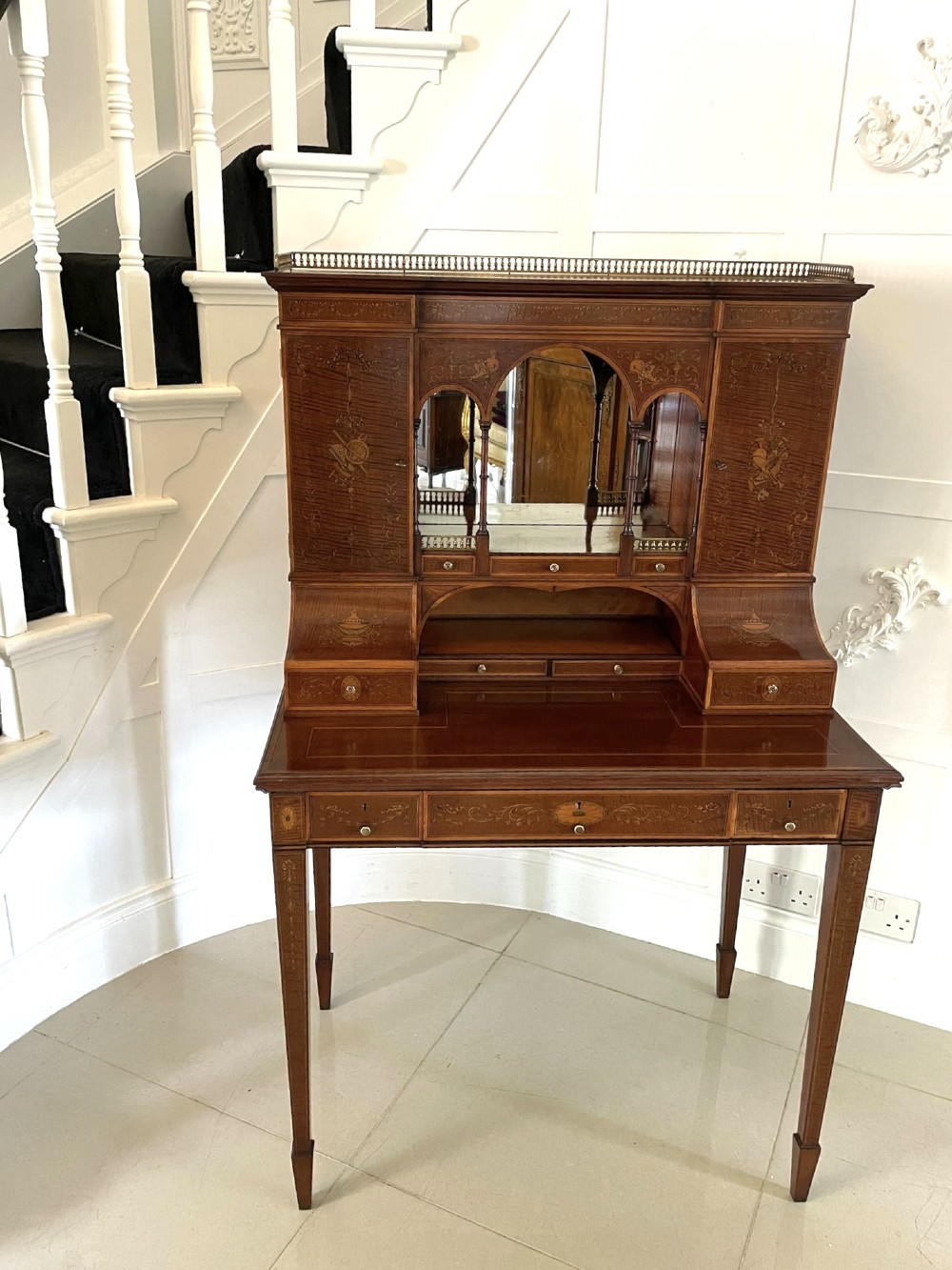 A Victorian inlaid mahogany writing desk featuring a brass gallery top, mirrored arched niche, floral marquetry cabinets, small drawers, and tapered square legs. View XV