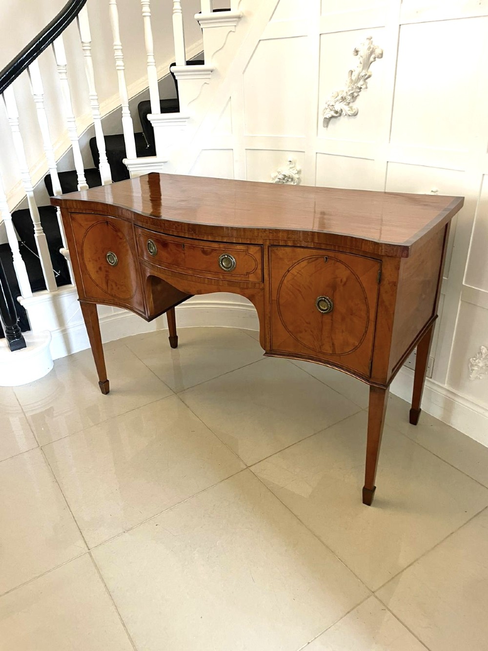 An antique satinwood serpentine-front sideboard featuring oval inlay panels, brass ring handles, and tapered square legs. View IV