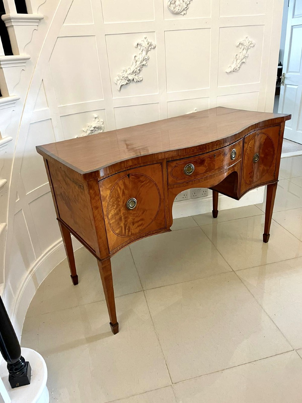 An antique satinwood serpentine-front sideboard featuring oval inlay panels, brass ring handles, and tapered square legs. View V