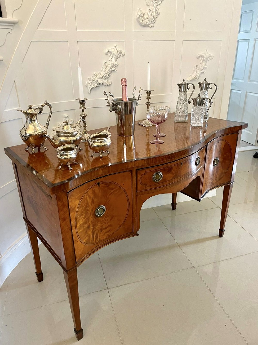 An antique satinwood serpentine-front sideboard featuring oval inlay panels, brass ring handles, and tapered square legs. View VI