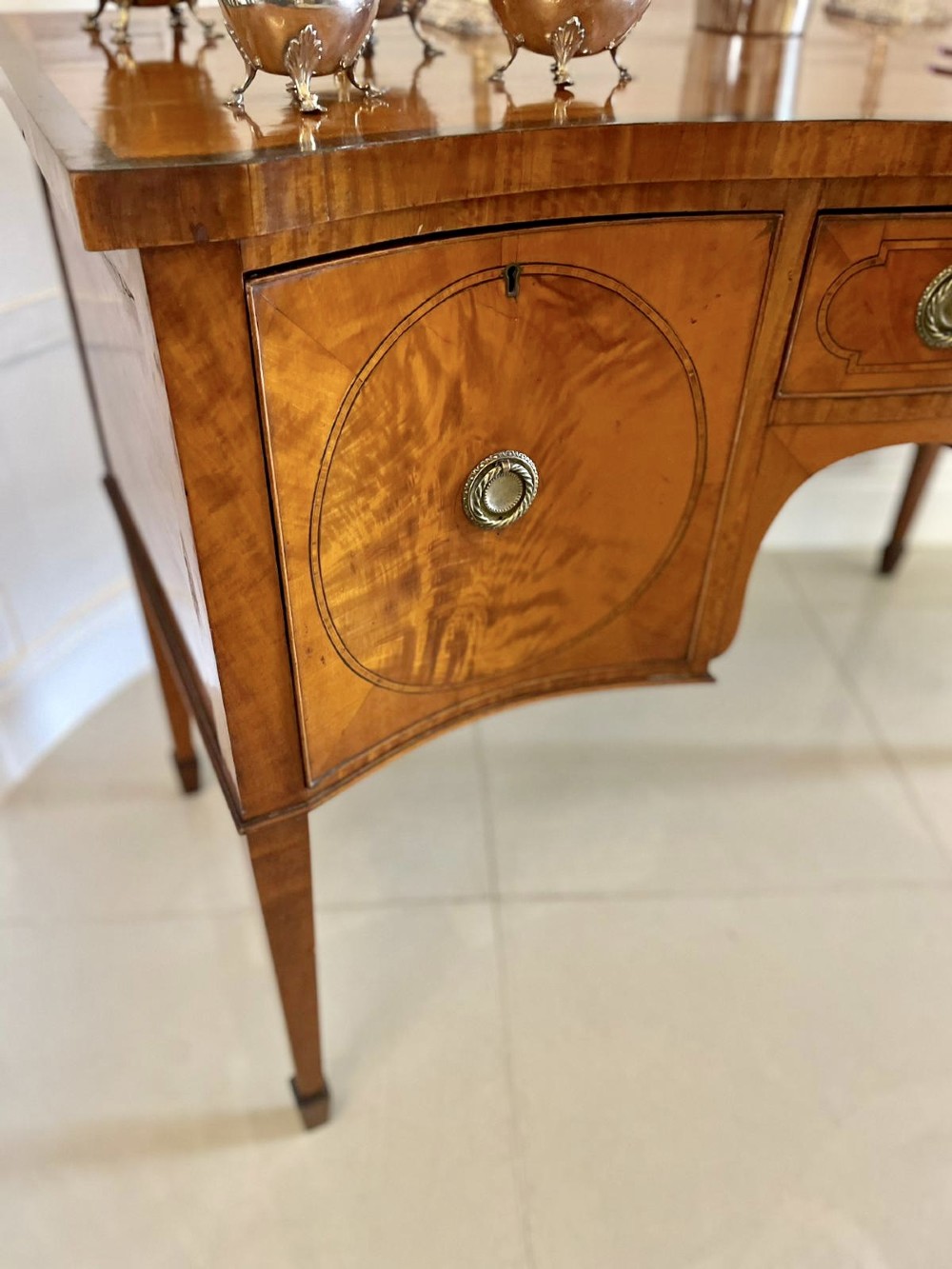An antique satinwood serpentine-front sideboard featuring oval inlay panels, brass ring handles, and tapered square legs. View XI