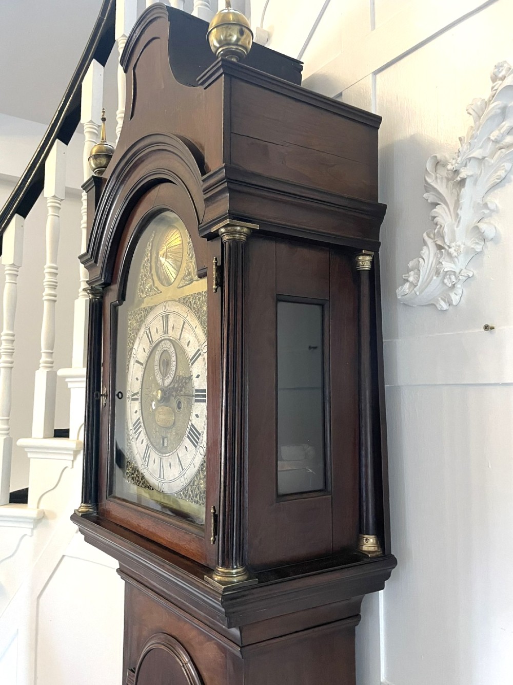 A George III mahogany longcase clock featuring an arched brass dial, urn finials, and an arched trunk door. View XI