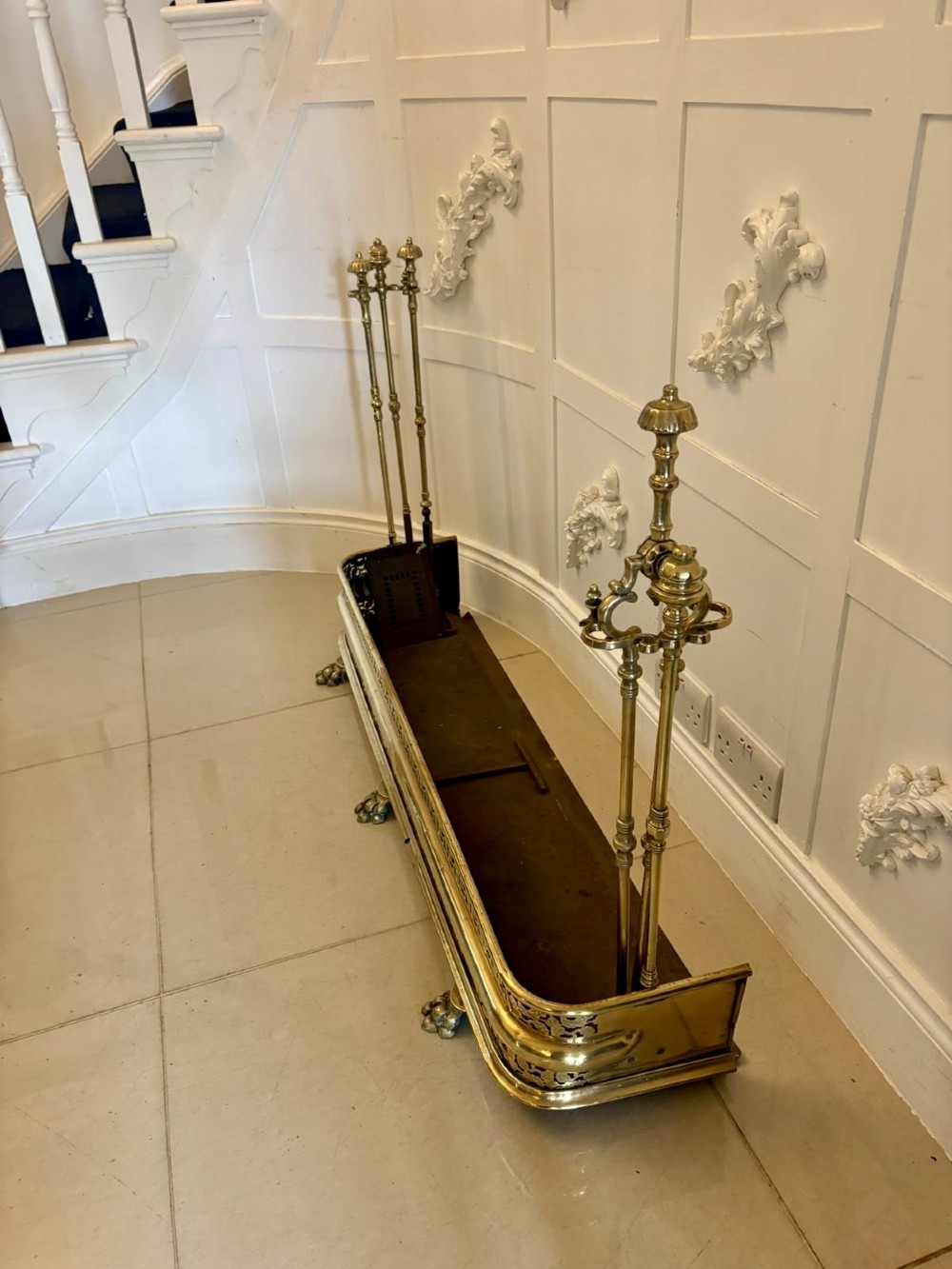 An antique Regency period brass fireplace fender featuring intricate pierced metalwork, claw feet, and matching original brass fire irons positioned at either end. View VIII
