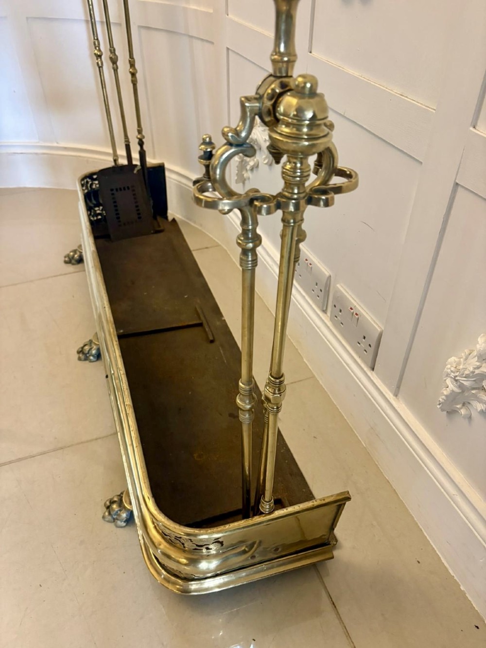 An antique Regency period brass fireplace fender featuring intricate pierced metalwork, claw feet, and matching original brass fire irons positioned at either end. View IX