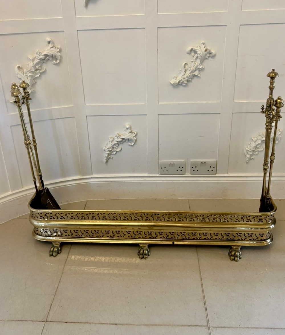 An antique Regency period brass fireplace fender featuring intricate pierced metalwork, claw feet, and matching original brass fire irons positioned at either end.