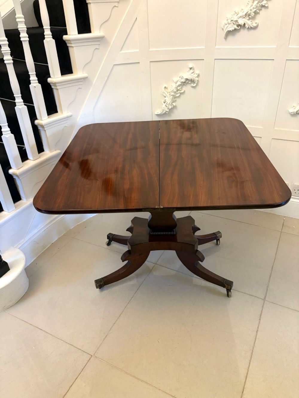 A Regency figured mahogany tea table with a fold-over rectangular top, central pedestal support, and four curved splayed legs on brass castors. View II