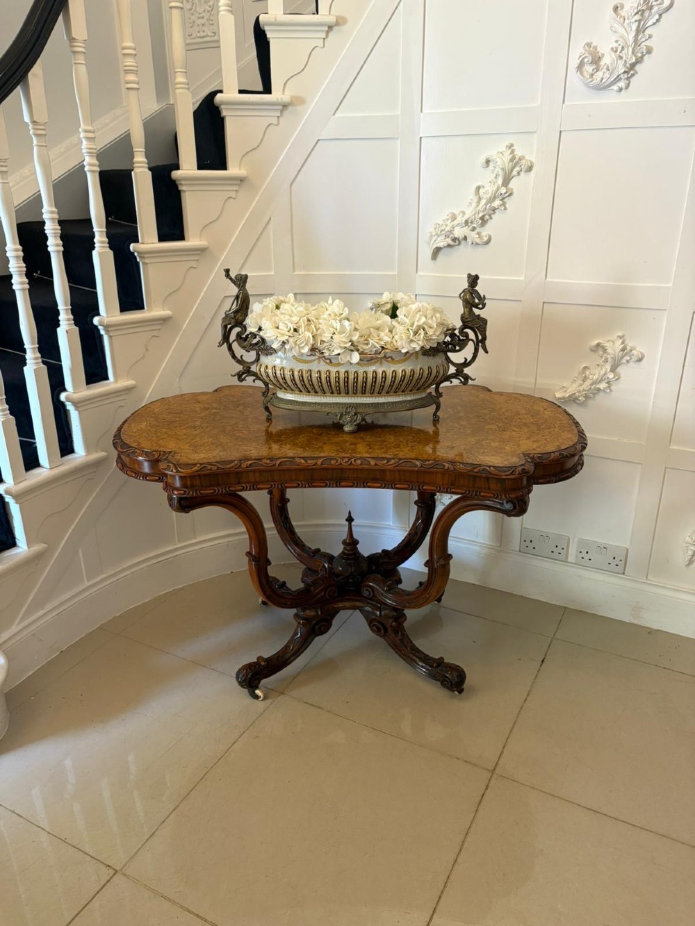 A Victorian burr walnut centre table featuring a shaped top, carved scroll frieze, and an elaborate four-legged base with a central turned finial. View II