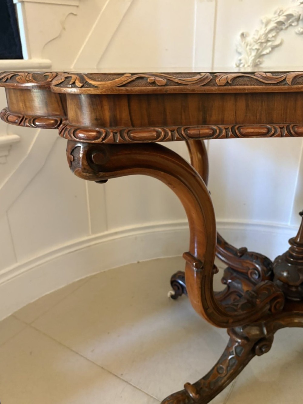 A Victorian burr walnut centre table featuring a shaped top, carved scroll frieze, and an elaborate four-legged base with a central turned finial. View V