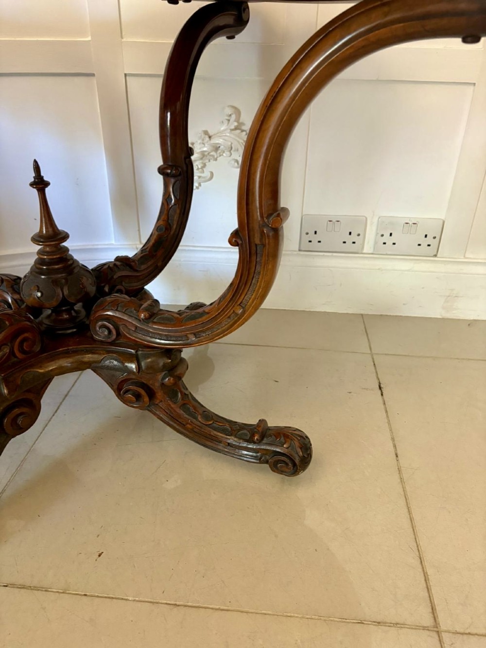 A Victorian burr walnut centre table featuring a shaped top, carved scroll frieze, and an elaborate four-legged base with a central turned finial. View X