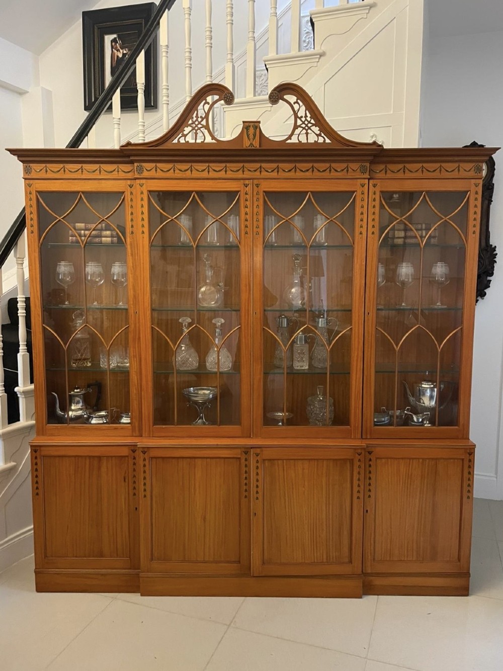 A large satinwood breakfront display cabinet featuring a swan-neck pediment, astragal glazed upper doors, and painted neoclassical motifs. View IV