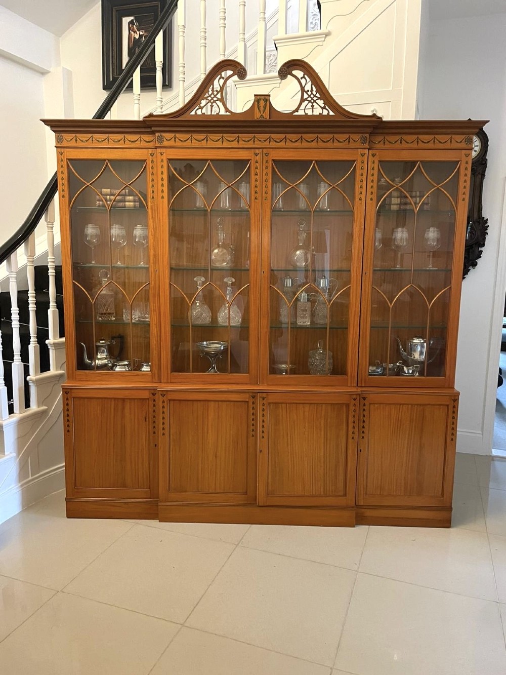 A large satinwood breakfront display cabinet featuring a swan-neck pediment, astragal glazed upper doors, and painted neoclassical motifs. View VI