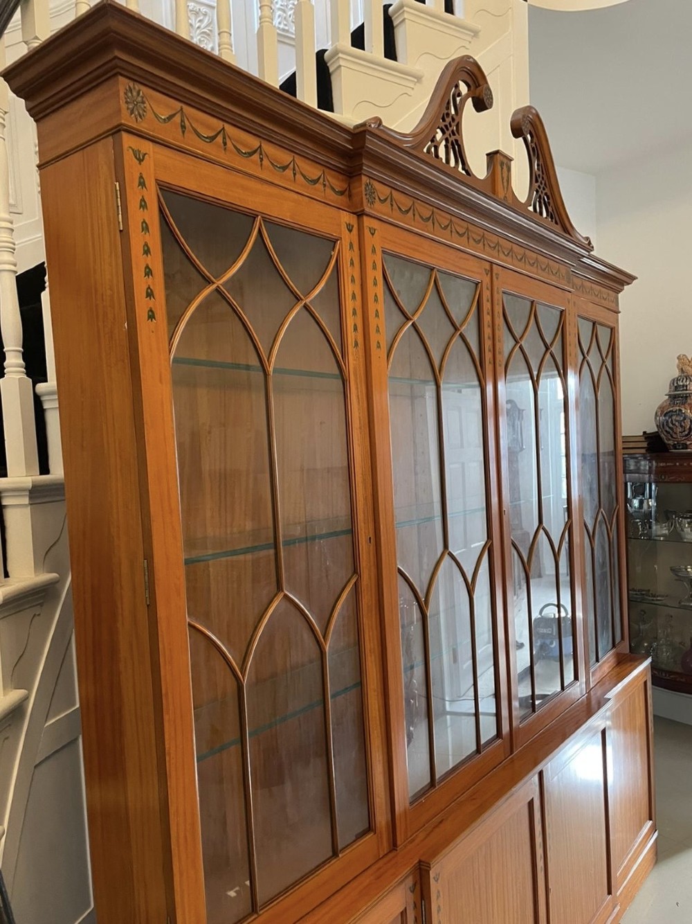 A large satinwood breakfront display cabinet featuring a swan-neck pediment, astragal glazed upper doors, and painted neoclassical motifs. View VII