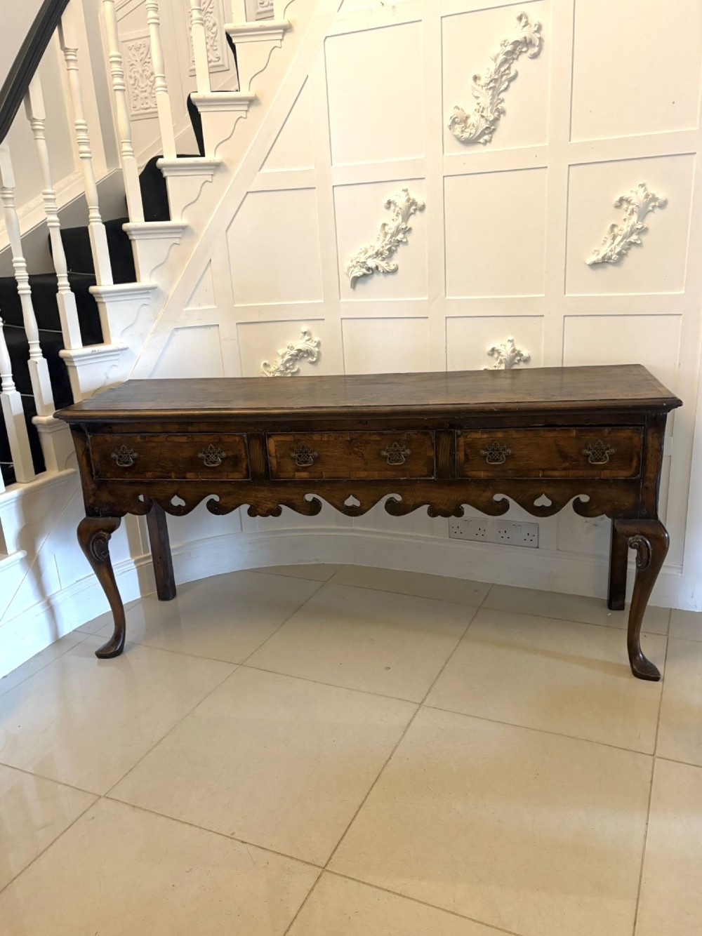 A dark brown oak Georgian style dresser base featuring three drawers with brass drop handles, a decorative scalloped apron, and front cabriole legs.