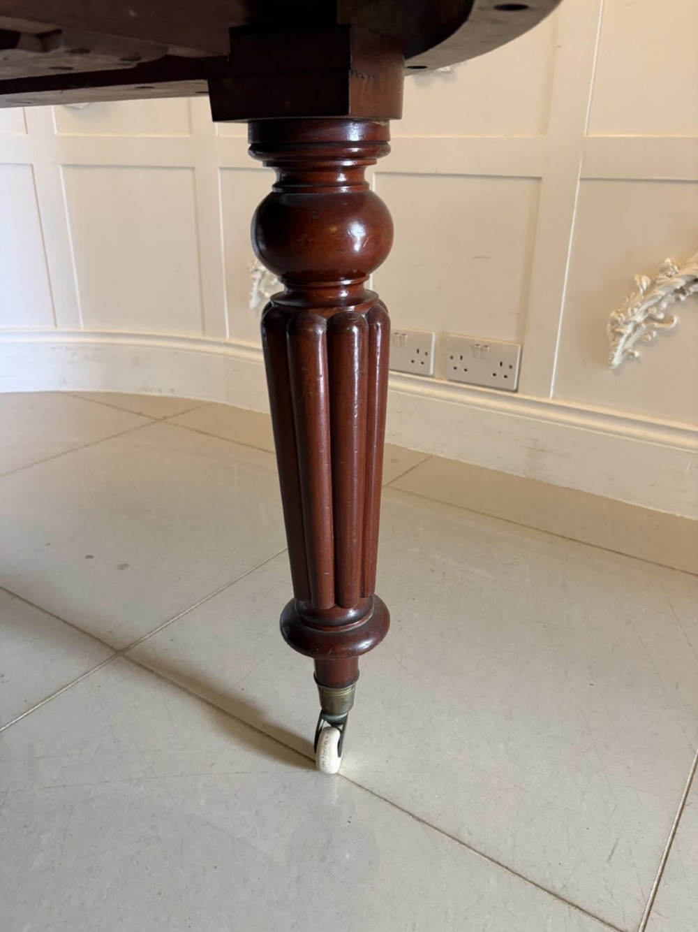 An antique Victorian oval mahogany dining table featuring a wind-out extending mechanism, fluted turned legs, and ceramic castors. View VII