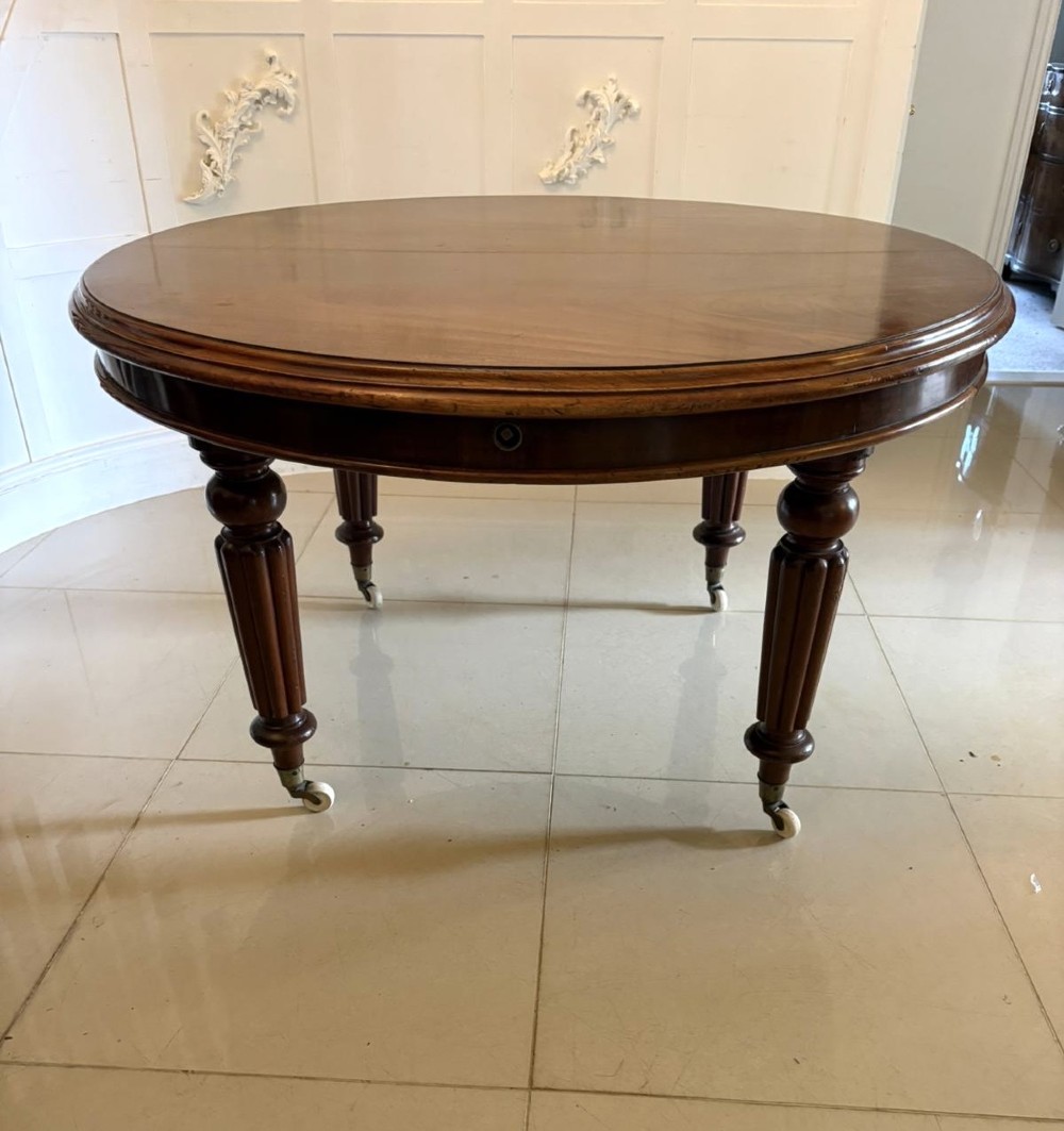 An antique Victorian oval mahogany dining table featuring a wind-out extending mechanism, fluted turned legs, and ceramic castors. View IX