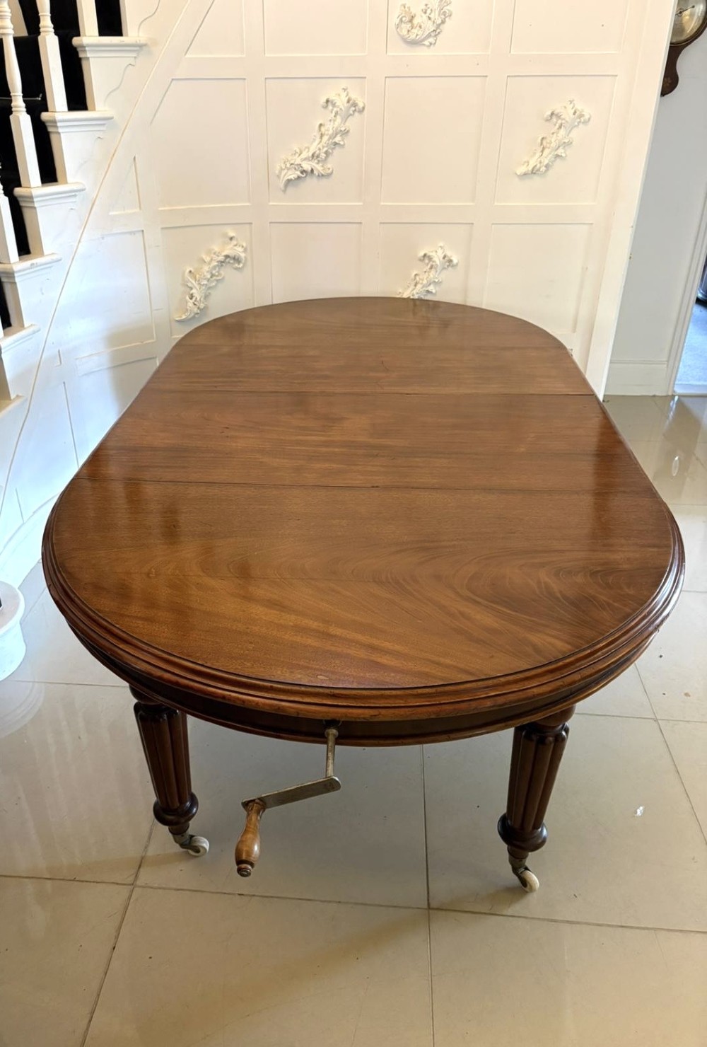 An antique Victorian oval mahogany dining table featuring a wind-out extending mechanism, fluted turned legs, and ceramic castors. View XI